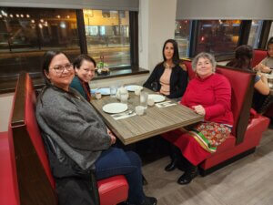 Four Indigenous women sit at a booth in a diner, facing the camera and smiling.